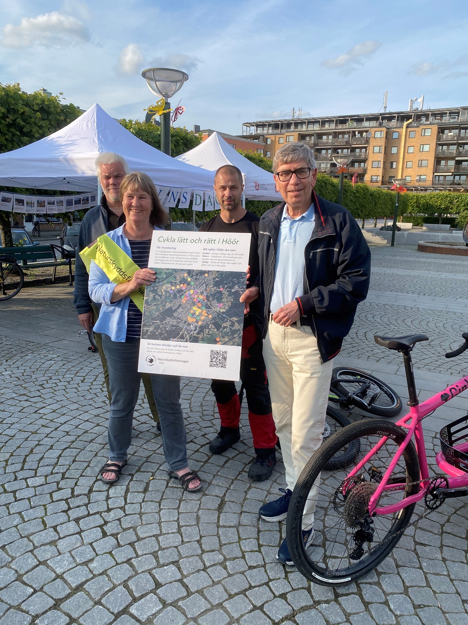 Fyra personer i torgmiljö, varav två håller i en affisch eller plakat. Bredvid står cyklar.