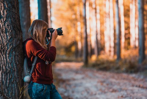 En blond kvinna med röd tröja och blå jeans fotograferar i skogen med en systemkamera. 