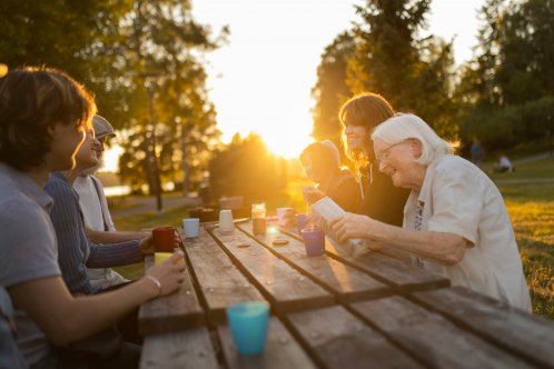 Sex personer sitter runt ett bord utomhus i naturen. En kvinna häller mjölk i en kaffekopp.