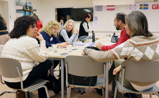 Flera personer sitter och pratar med varandra runt ett bord i ett upplyst klassrum.