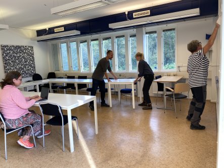 Två personer flyttar bord, en person rengör en whiteboardtavla och en person sitter vid en bärbar dator.