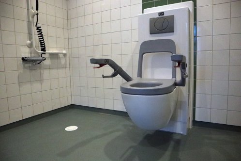 En handikappanpassad toalett i ett utrymme med kaklade väggar och plastmatta på golvet.