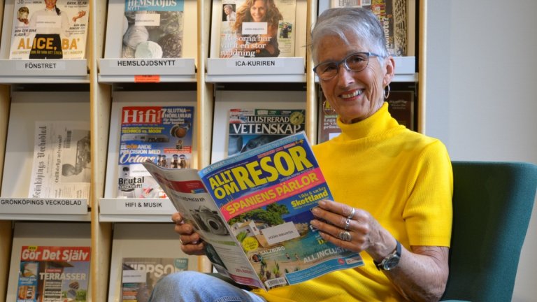 En äldre, kortklippt kvinna sitter och håller i tidningen Allt om resor. Hon har en gul polojumper på sig och ler in i kameran. I hyllan bakom henne syns rader med tidskrifter.