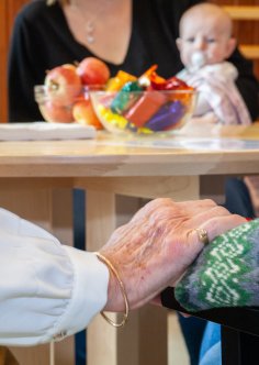 En äldre person har lagt sin hand på en annan persons arm på ett omhändertagande och stöttande sätt. I bakgrunden syns ett bord.