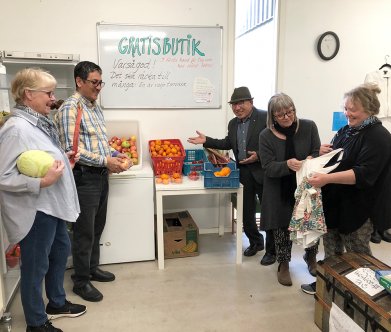 Fem personer står i en lokal. De håller i grönsaker och på väggen sitter en skylt där det står "gratisbutik".