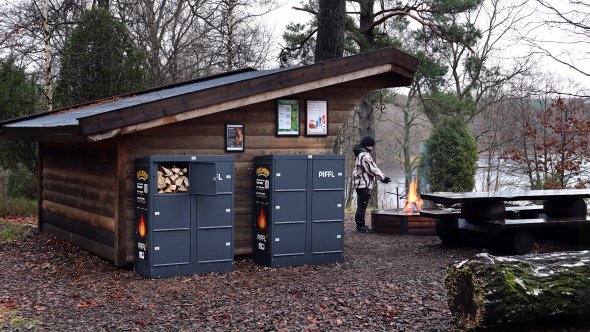 En slogbod i skogen, i bakgrunden syns en glimt av Vaxsjön. En person står framför slogboden och grillar på en öppen eld. På sidan av boden två skåp, där inne syns ved.