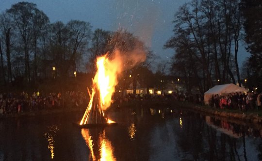 En mörk bild med en sjö, i mitten brinner det en stor eld. På kanten runt om sjön står en folkmassa. Ett vit partytält syns också.